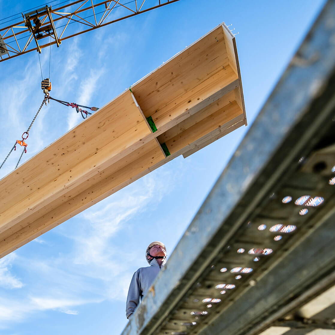 Holzbauelement wird mit einem Kran angehoben, während eine Person auf der Baustelle nach oben blickt.