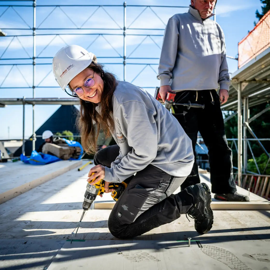 Mitarbeiterin verschraubt ein Bauteil auf einer Baustelle, weitere Teammitglieder und Gerüstkonstruktion im Hintergrund.