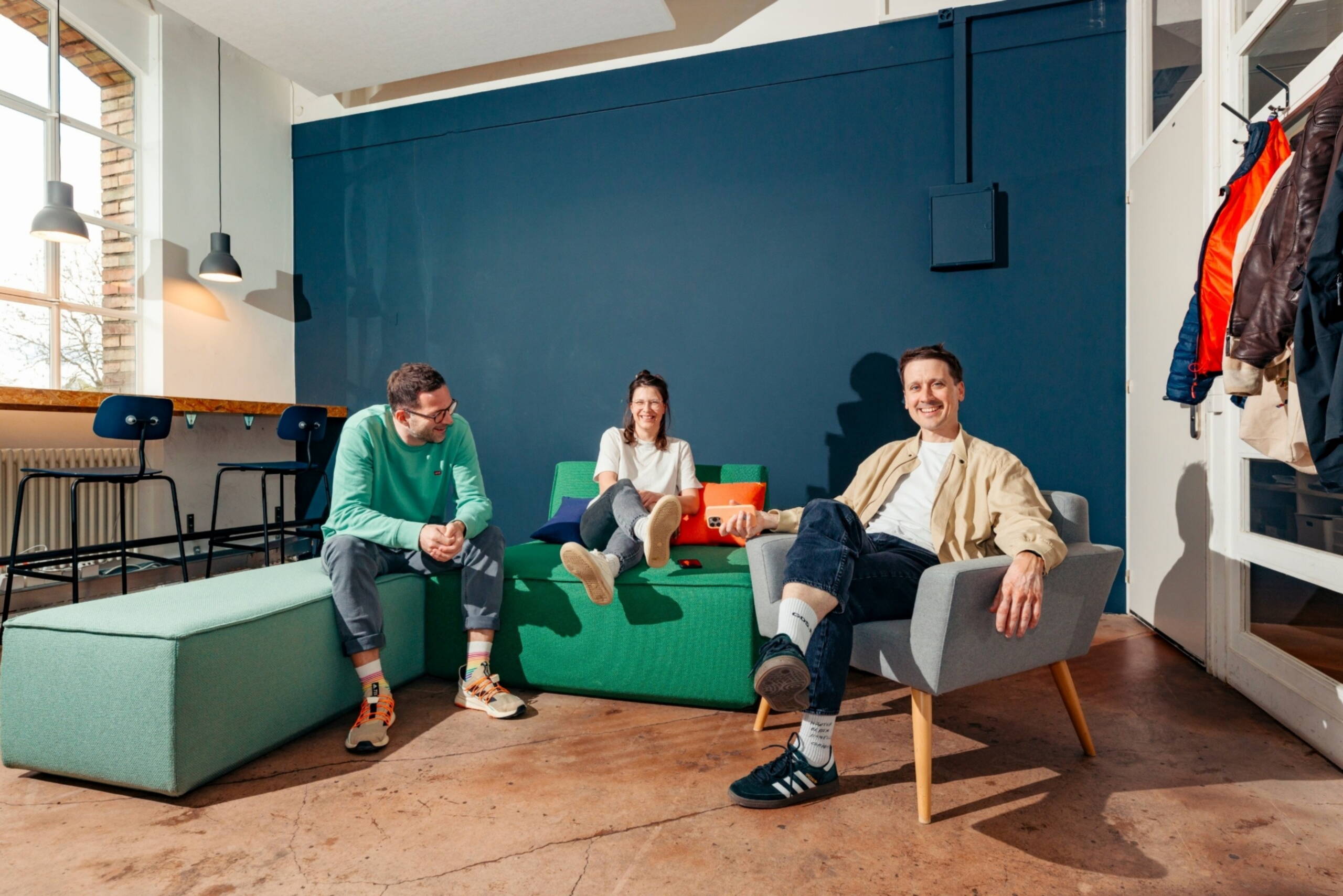 Team Drei Personen (Chrigi, Isa, Tobi) sitzen entspannt auf Sofas und einem Sessel in einem hellen Büro mit dunkelblauer Wand.